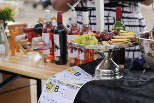 A table at a business expo displays wine bottles, a wine glass, and a platter with grapes and crackers. A festive, elegant atmosphere is conveyed.