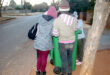 A group of people stand around a green garbage bin on a quiet suburban street. They're bundled in winter clothing, creating a sense of community and cooperation.