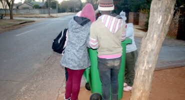 A group of people stand around a green garbage bin on a quiet suburban street. They're bundled in winter clothing, creating a sense of community and cooperation.
