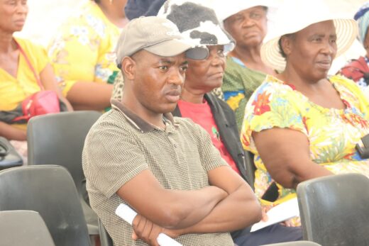A man with a cap sits cross-armed in a crowd, appearing focused. Behind him, elderly women in colorful attire and hats listen attentively. Community event.