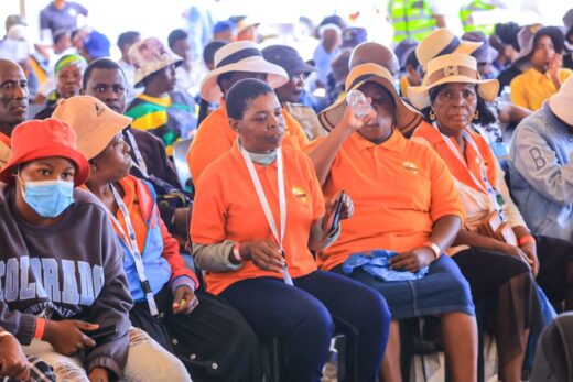 A diverse group of people, mostly women, sit together wearing orange shirts and hats. Some drink water, creating a relaxed and attentive atmosphere.