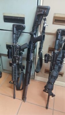 Rifles and a shotgun with tactical gear lean against a cabinet on a tiled floor. The scene conveys a sense of readiness and security.