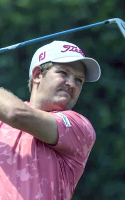 Golfer in a pink shirt and white cap swings a club, focused expression, green blurred background. Concentrated action during a game.