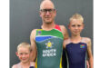 A man wearing a South Africa sports uniform stands smiling between two young boys, both in matching athletic outfits with South African flags, against a gray background.