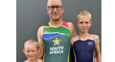 A man wearing a South Africa sports uniform stands smiling between two young boys, both in matching athletic outfits with South African flags, against a gray background.