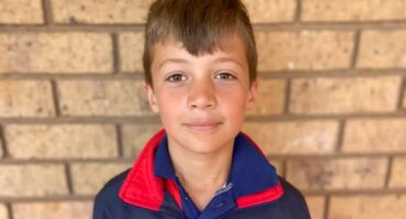 A young boy with short brown hair stands against a brick wall, wearing a blue jacket with a red collar. He has a calm expression.