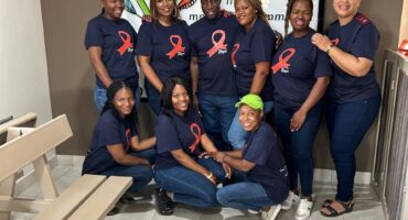 A group of eight people, smiling, pose in a room with beige benches. All wear navy shirts with red awareness ribbons. The atmosphere is friendly and supportive.