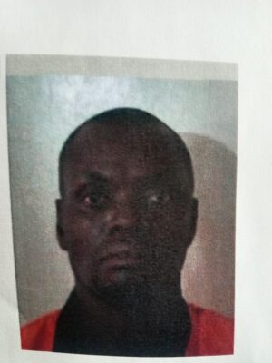 A low-quality photo shows a person with a serious expression against a neutral background. They are wearing a dark shirt with a red garment over it.