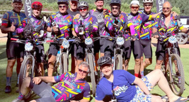 A group of smiling cyclists in colorful jerseys and helmets posing with their bikes on a sunny day. Two people relax in front, creating a joyful atmosphere.