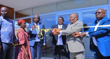 A group of six people, including three men in suits and a woman in a red patterned dress, are at a ribbon-cutting ceremony in front of a building. The mood is celebratory.