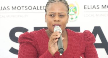 A person in a red textured jacket speaks into a microphone. A backdrop with "Elias Motsoaledi Local Municipality" is partially visible. The tone is serious.