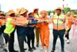 A group of people in bright orange safety outfits and hats cut a red ribbon outdoors, celebrating an event. They appear joyful and united.