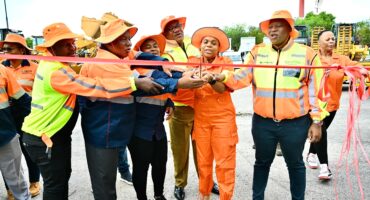 A group of people in bright orange safety outfits and hats cut a red ribbon outdoors, celebrating an event. They appear joyful and united.