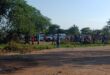 A large group of people gathers near parked vehicles on a dirt road under a clear blue sky. Trees frame the scene, evoking a communal atmosphere.