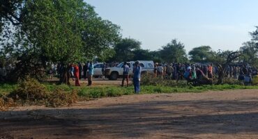 A large group of people gathers near parked vehicles on a dirt road under a clear blue sky. Trees frame the scene, evoking a communal atmosphere.