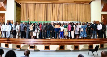 A large group of people stands on a wooden stage in an auditorium, smiling at the camera. The background features beige curtains and wooden decor.