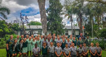 A group of school children in uniforms stand in rows on a lush green lawn, smiling. Trees and a white building with a flag in the background.