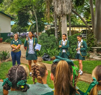 A group of girls in green uniforms stands outdoors. One speaks animatedly, holding a paper, as others listen. The scene is lively with lush greenery.