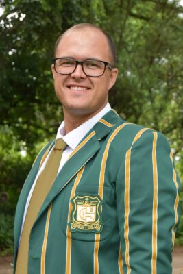 A smiling man in glasses wearing a green and yellow-striped blazer with a crest stands outside. Lush greenery forms the bright, vibrant background.