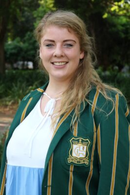 Smiling woman with long blonde hair, wearing a green blazer with yellow stripes and an emblem over a light blue and white top, outdoors with greenery.