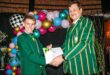 Two people in green-striped blazers smile as they exchange a certificate. Colorful balloons form an arch in the background, adding a festive mood.
