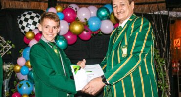 Two people in green-striped blazers smile as they exchange a certificate. Colorful balloons form an arch in the background, adding a festive mood.
