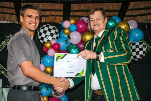 Two men smiling and shaking hands at an award ceremony. One holds a certificate. Colorful balloons form a festive backdrop, conveying celebration.