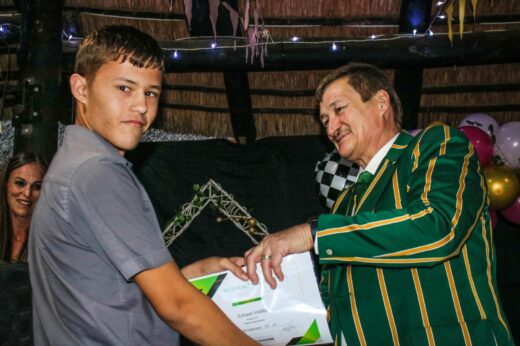 A young man in a gray shirt receives a certificate from a man in a green striped blazer at an indoor ceremony, with festive decorations around.