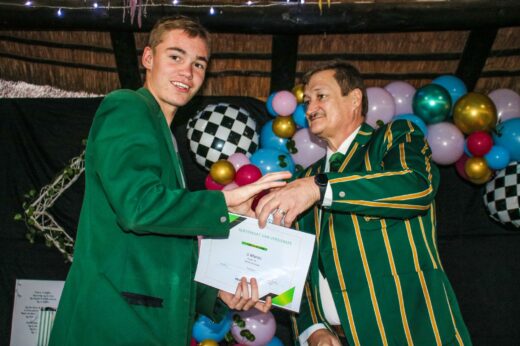 A young man in a green blazer receives a certificate from an older man in a striped blazer. They stand in front of a festive balloon display, smiling.