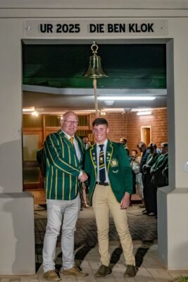 Two men in green blazers with yellow stripes shake hands under a sign that reads "UR 2025 DIE BEN KLOK." A crowd watches in the background.