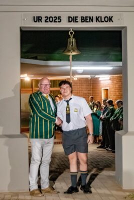 Two men stand under a sign reading "UR 2025 DIE BEN KLOK," shaking hands. One wears a green and yellow striped jacket, the other in school uniform. A bell hangs above.