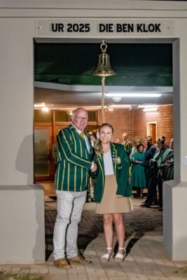 A man and a young woman, both in green blazers, stand smiling and shaking hands under a bell. A small group watches from a well-lit room behind.