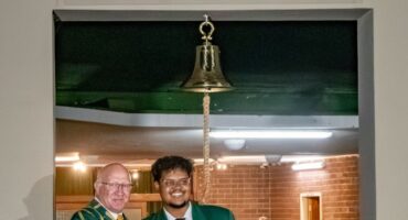 Two men in green blazers smiling under a bell with "UR 2025 DIE BEN KLOK" sign. They stand in a warmly lit room with people in the background.