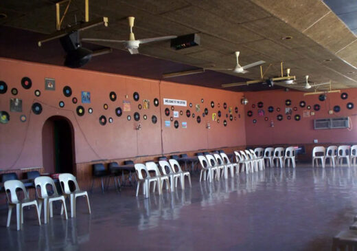 Spacious event hall with vinyl records decorating one wall, surrounded by white plastic chairs. Ceiling fans hang overhead, creating a retro ambiance.
