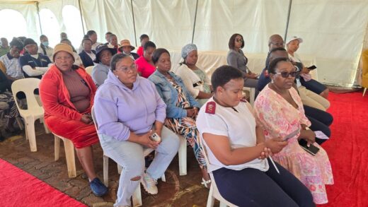 A diverse group of people sits attentively on white chairs inside a tent with a red carpet. Some hold phones, projecting a serious, focused mood.