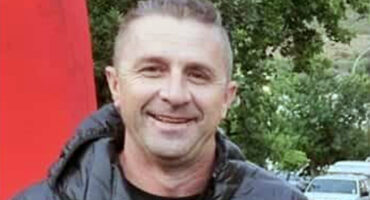 A man in a black jacket smiles warmly, standing outdoors against a backdrop of green trees and parked cars, conveying a friendly and relaxed mood.