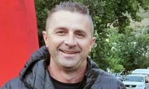 A man in a black jacket smiles warmly, standing outdoors against a backdrop of green trees and parked cars, conveying a friendly and relaxed mood.