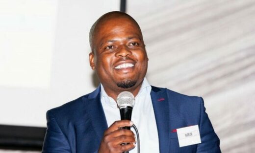 A smiling man in a blue suit holds a microphone, standing against a light background. He appears confident and is possibly speaking at an event.