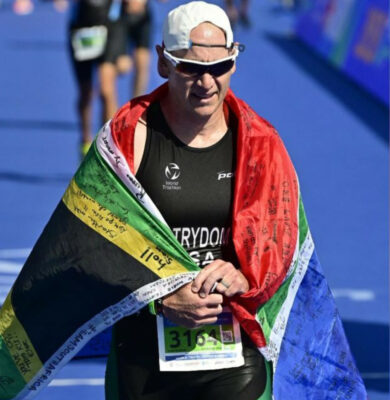Athlete in black triathlon gear and white cap, wrapped in a flag, finishes a race on a blue track. The flag has messages on it, showing triumph and pride.