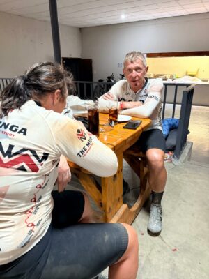 Two cyclists in matching jerseys sit at a wooden table, appearing tired. Drinks and phones are on the table, creating a relaxed post-ride atmosphere.