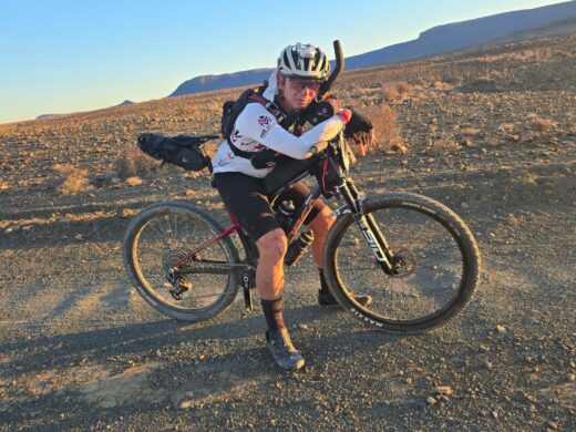 Cyclist rests on a bike in a rocky desert landscape at sunset, wearing a white jersey and helmet, conveying determination and exhaustion.