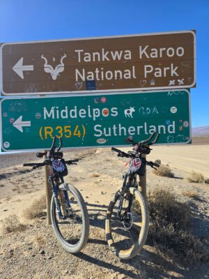 Two bicycles lean against signs for Tankwa Karoo National Park and Middelpos/Sutherland (R354) on a dirt road under a clear blue sky.