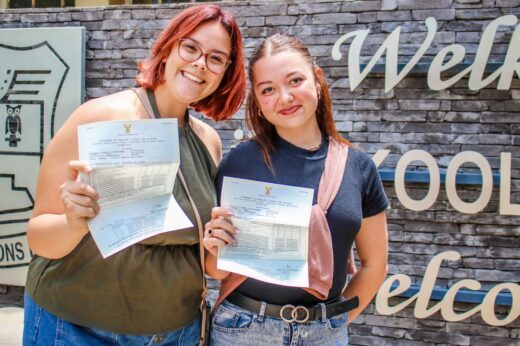 Two women smile, holding up certificates in front of a stone wall with partial words visible. They appear happy, celebrating achievements.