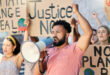 Protesters at a rally, with a man in a pink shirt passionately speaking through a megaphone. Signs read "Justice Now" and "No Plan B."