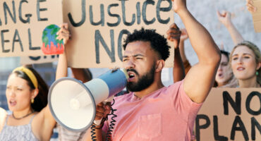 Protesters at a rally, with a man in a pink shirt passionately speaking through a megaphone. Signs read "Justice Now" and "No Plan B."