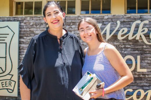 A smiling woman in a black blouse and a young woman in glasses hold mail, standing in front of a stone wall with "Welcome" partially visible.