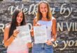 Two young women stand smiling, holding certificates against a brick wall with partially visible welcoming text. The mood is celebratory and proud.