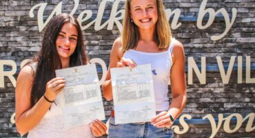 Two young women stand smiling, holding certificates against a brick wall with partially visible welcoming text. The mood is celebratory and proud.