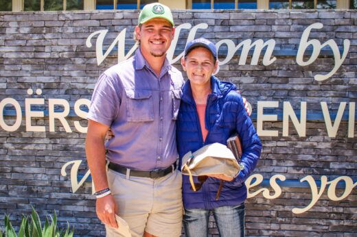 A smiling man and woman stand outdoors in front of a stone wall with text. The woman holds a brown package. Both wear casual attire and caps.