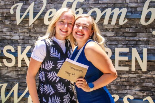 Two smiling young women pose in front of a stone wall with large text. One holds an envelope. Both appear joyful and celebratory.
